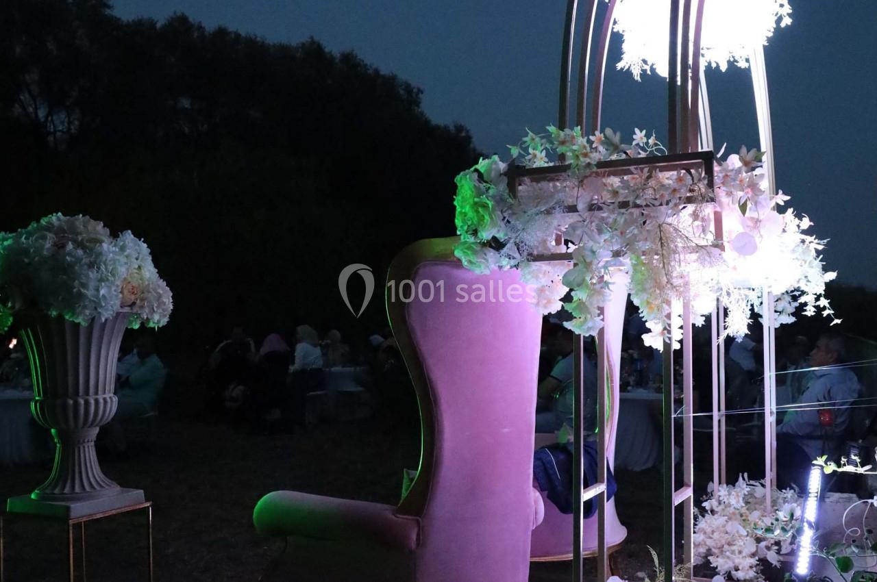 Fauteuil rose et arche décorée de fleurs blanches illuminées, installés en extérieur à la tombée de la nuit.