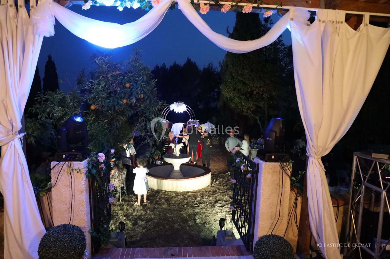 Vue nocturne d'une cérémonie en extérieur avec des invités, éclairages décoratifs et une arche fleurie au centre.