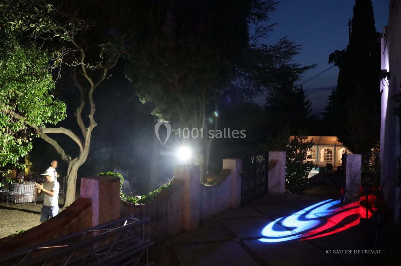 Un jardin éclairé de nuit avec des projections lumineuses colorées au sol et une personne marchant près des arbres.