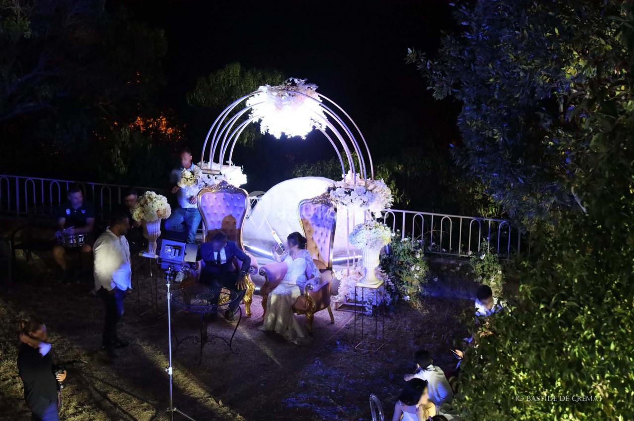 Un couple assis sur un canapé décoré, entouré de fleurs et éclairé, lors d'un événement en extérieur de nuit.