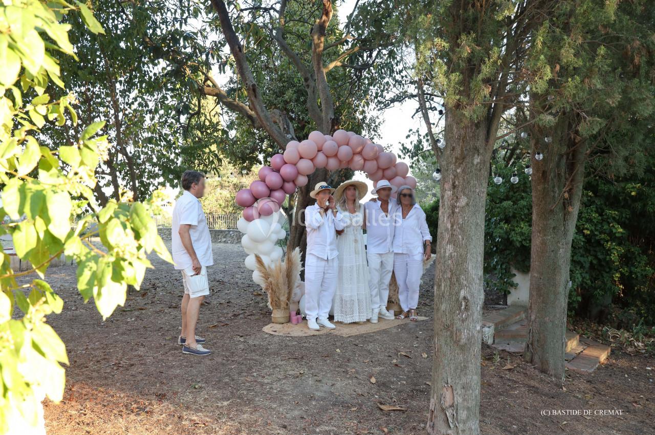 Un groupe de personnes en tenue blanche pose sous une arche décorée de ballons dans un jardin ombragé.