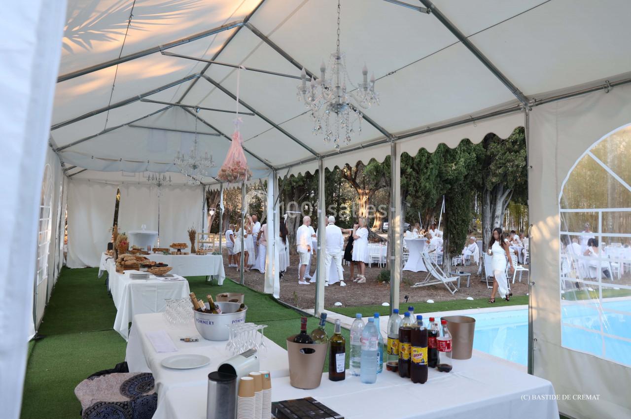 Intérieur d'une tente blanche aménagée pour un événement, avec tables dressées et invités en tenue blanche à l'extérieur.