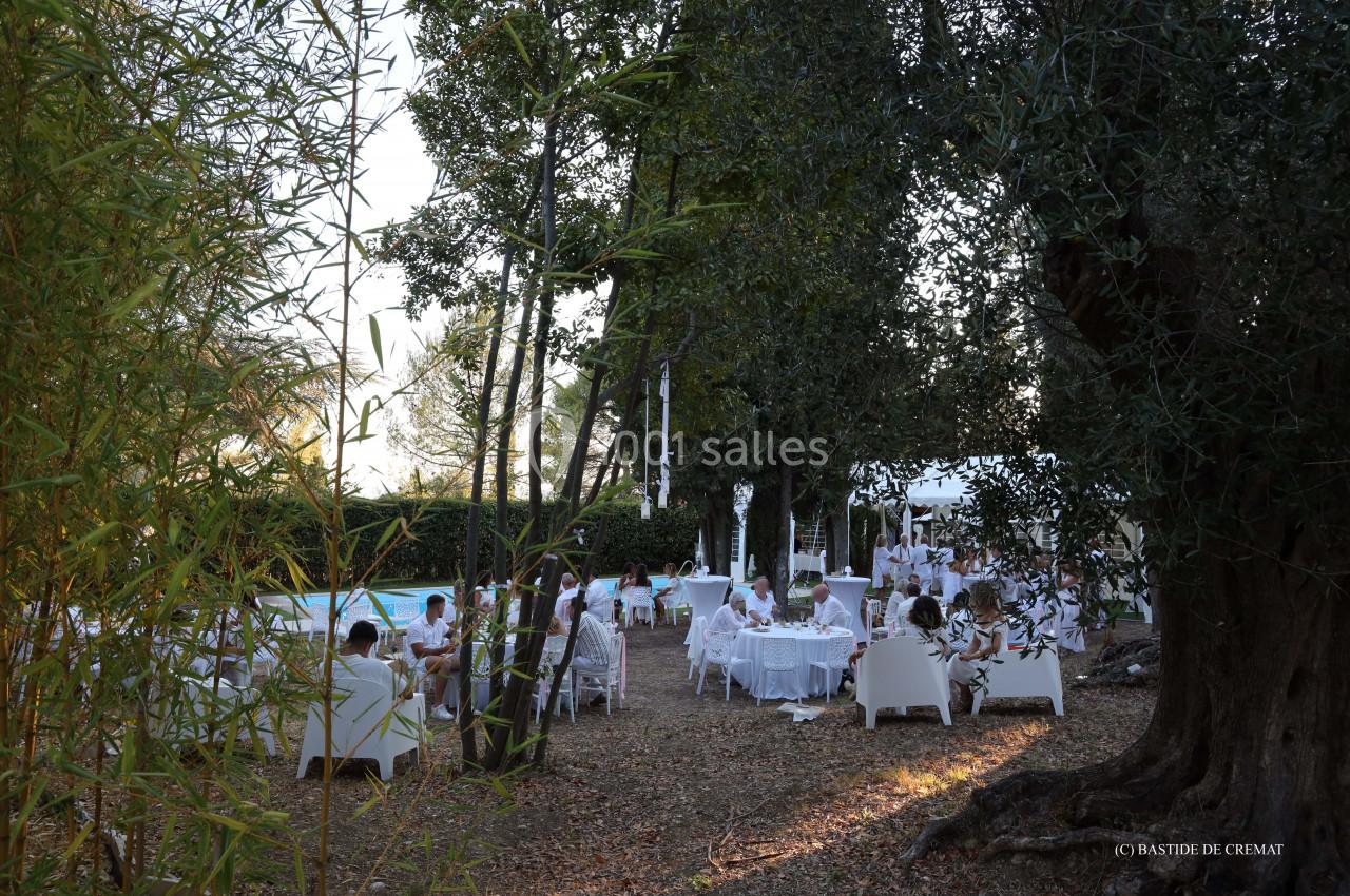 Des convives assis à des tables blanches dans un jardin ombragé, entouré d'arbres et de végétation.