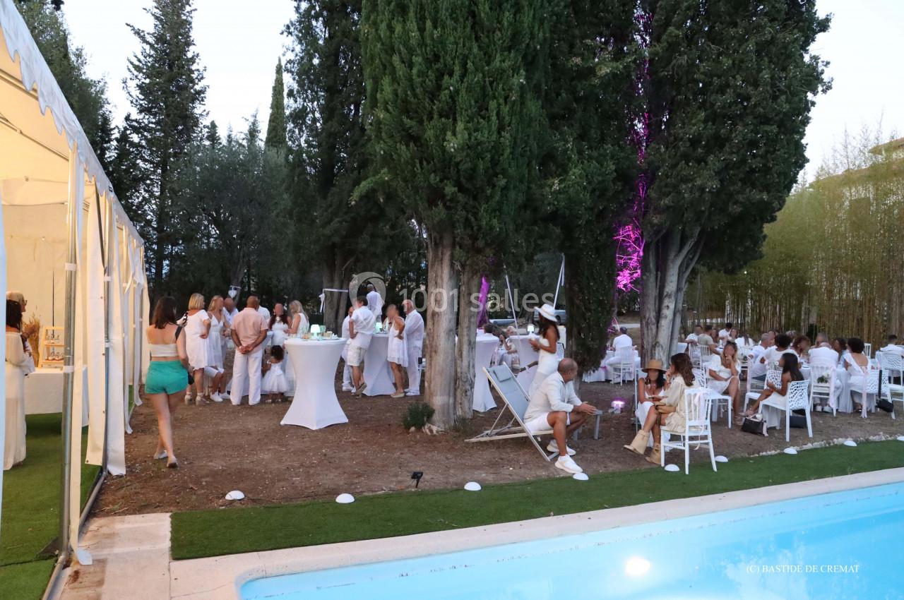 Groupe de personnes en tenue blanche réunies en extérieur près d'une piscine, entourées d'arbres et d'une tente éclairée.