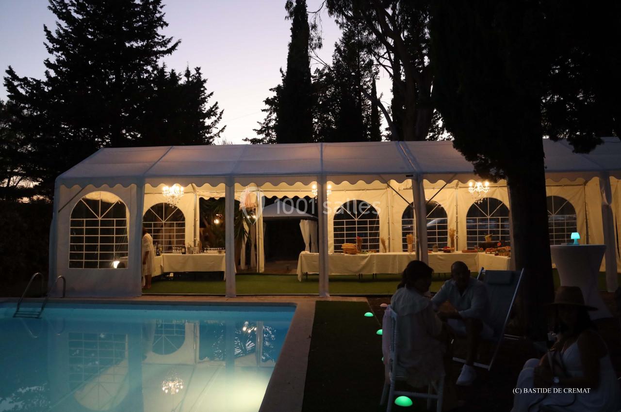 Tente éclairée en soirée près d'une piscine, entourée d'arbres et de personnes discutant à l'extérieur.