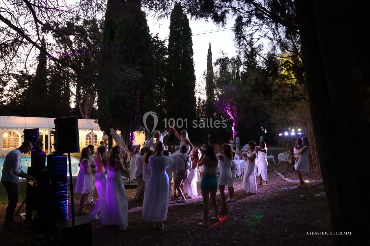 Des personnes vêtues de blanc dansent en plein air au crépuscule, entourées d'arbres et de lumières colorées.