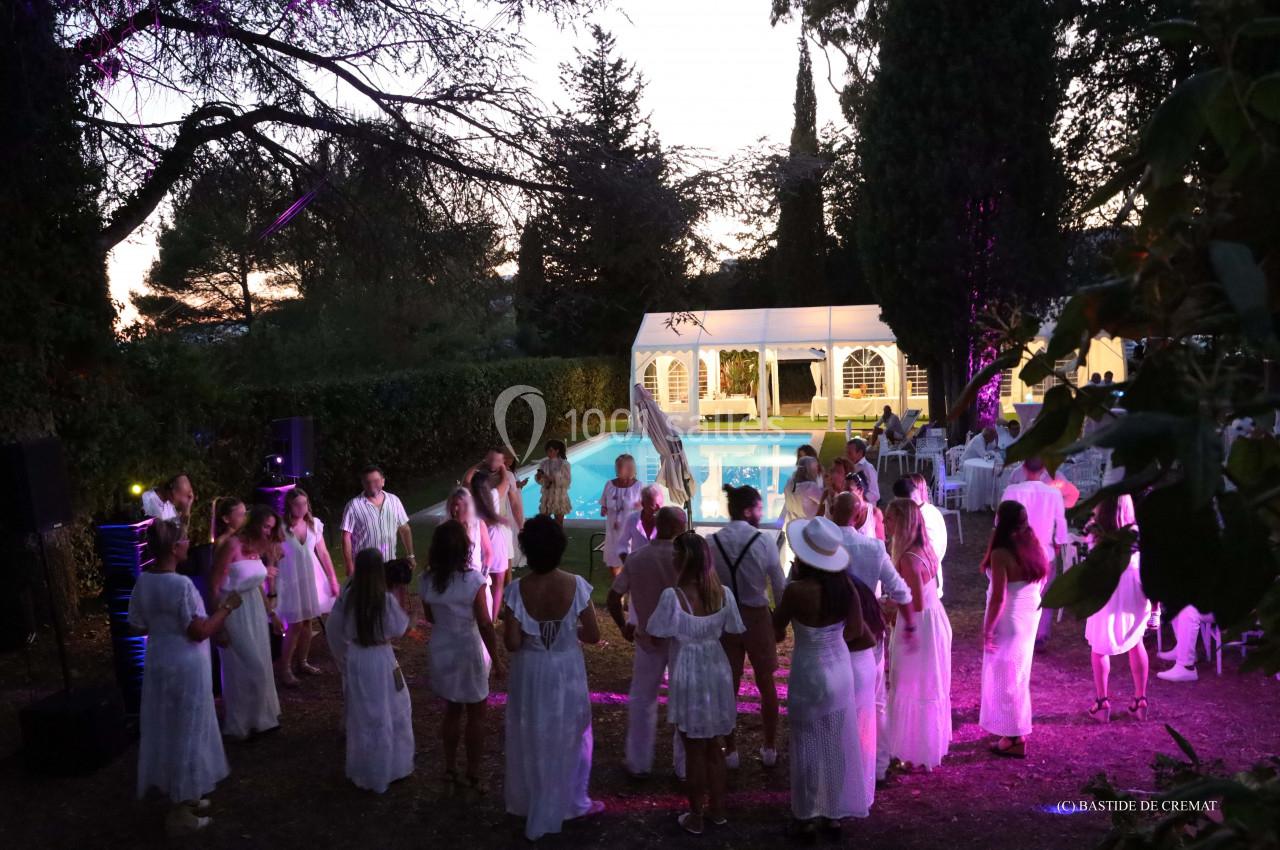 Groupe de personnes en tenue blanche dans un jardin, dansant près d'une piscine éclairée au crépuscule.