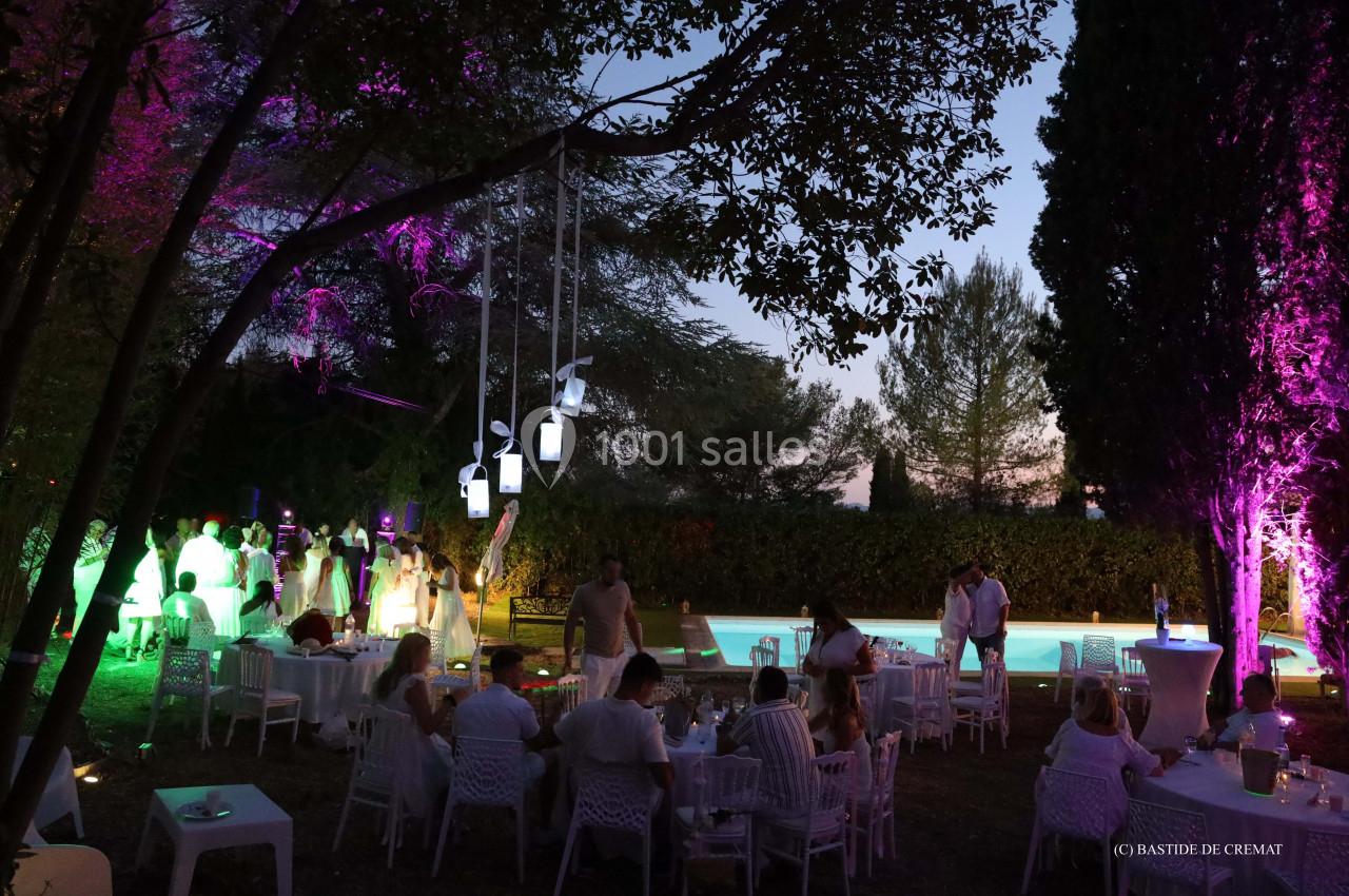 Soirée en plein air avec invités autour de tables blanches, éclairages colorés et piscine en arrière-plan.