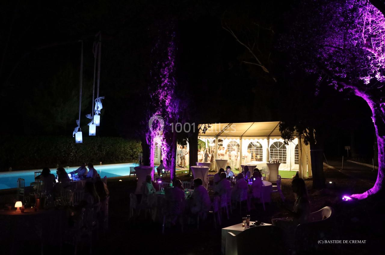 Vue nocturne d'une réception en plein air avec tente éclairée, tables dressées et éclairages colorés dans un jardin.