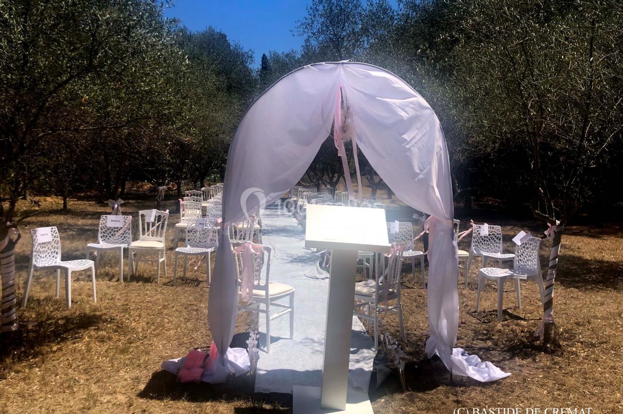Arche blanche décorée pour une cérémonie en plein air, entourée de chaises disposées dans un cadre naturel.