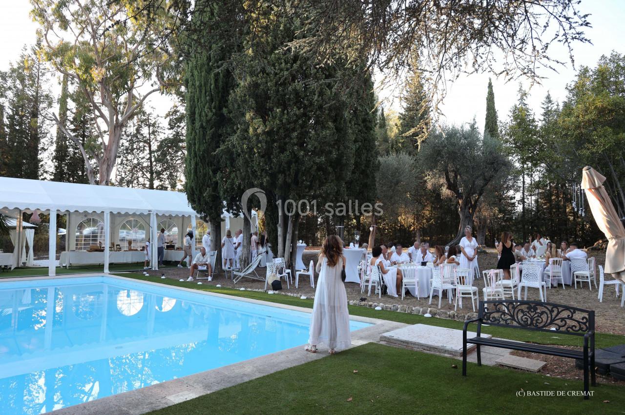 Des invités en tenue blanche sont rassemblés autour de tables près d'une piscine, entourés d'arbres et d'une tente blanche.