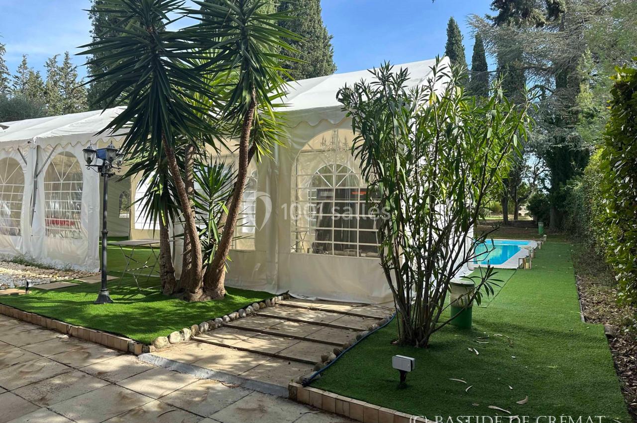 Tente blanche avec fenêtres cintrées, entourée de végétation, près d'une piscine et d'une allée dallée.