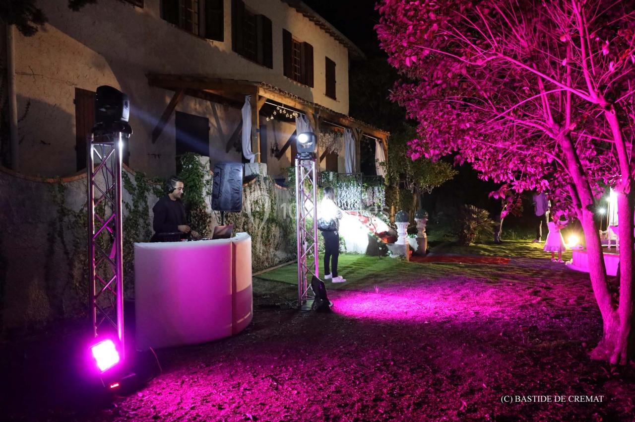 DJ jouant de la musique en extérieur, éclairé par des lumières roses, devant une bâtisse entourée de végétation.