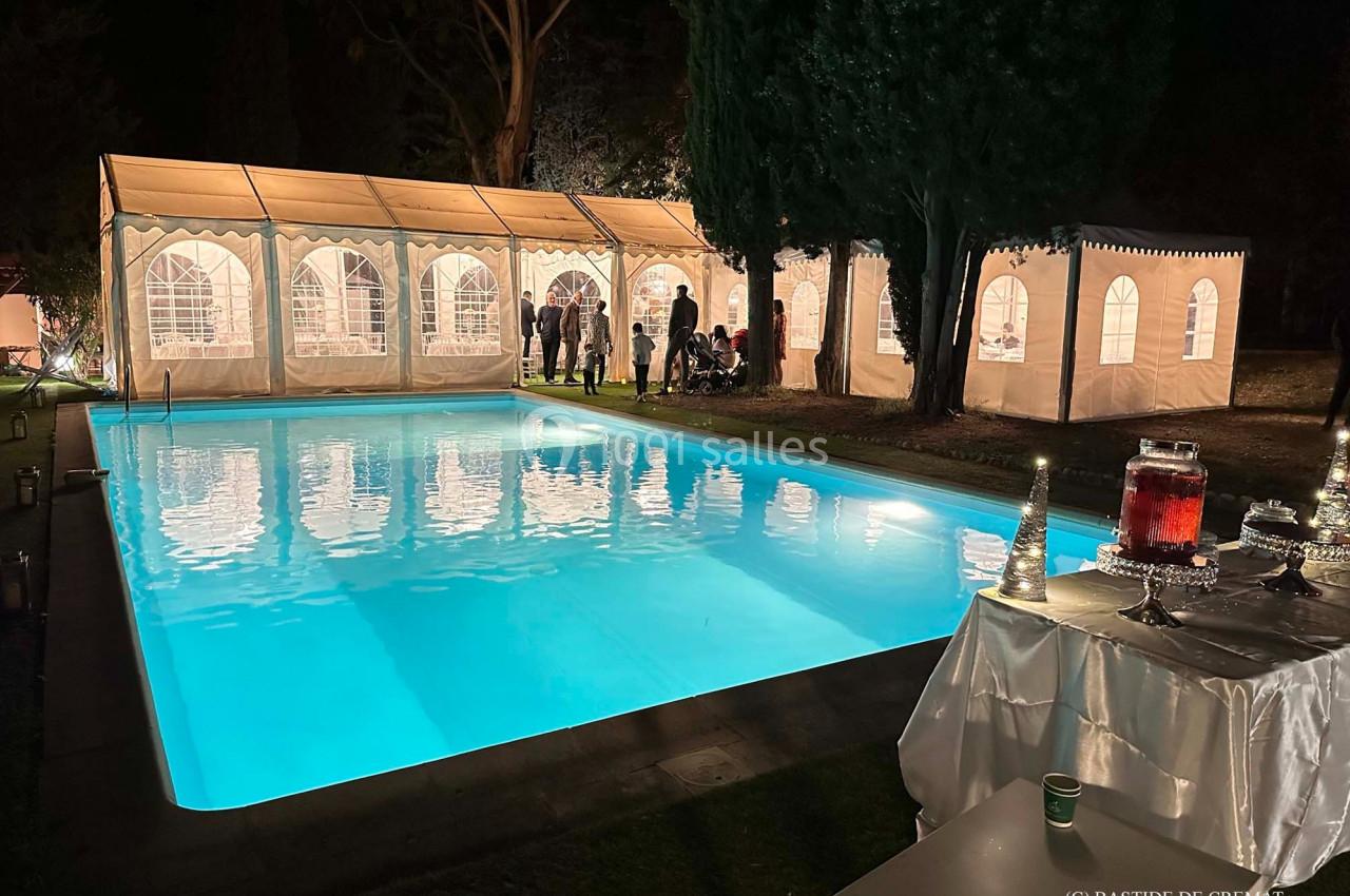 Piscine éclairée de nuit avec des tentes blanches illuminées en arrière-plan et une table dressée au premier plan.