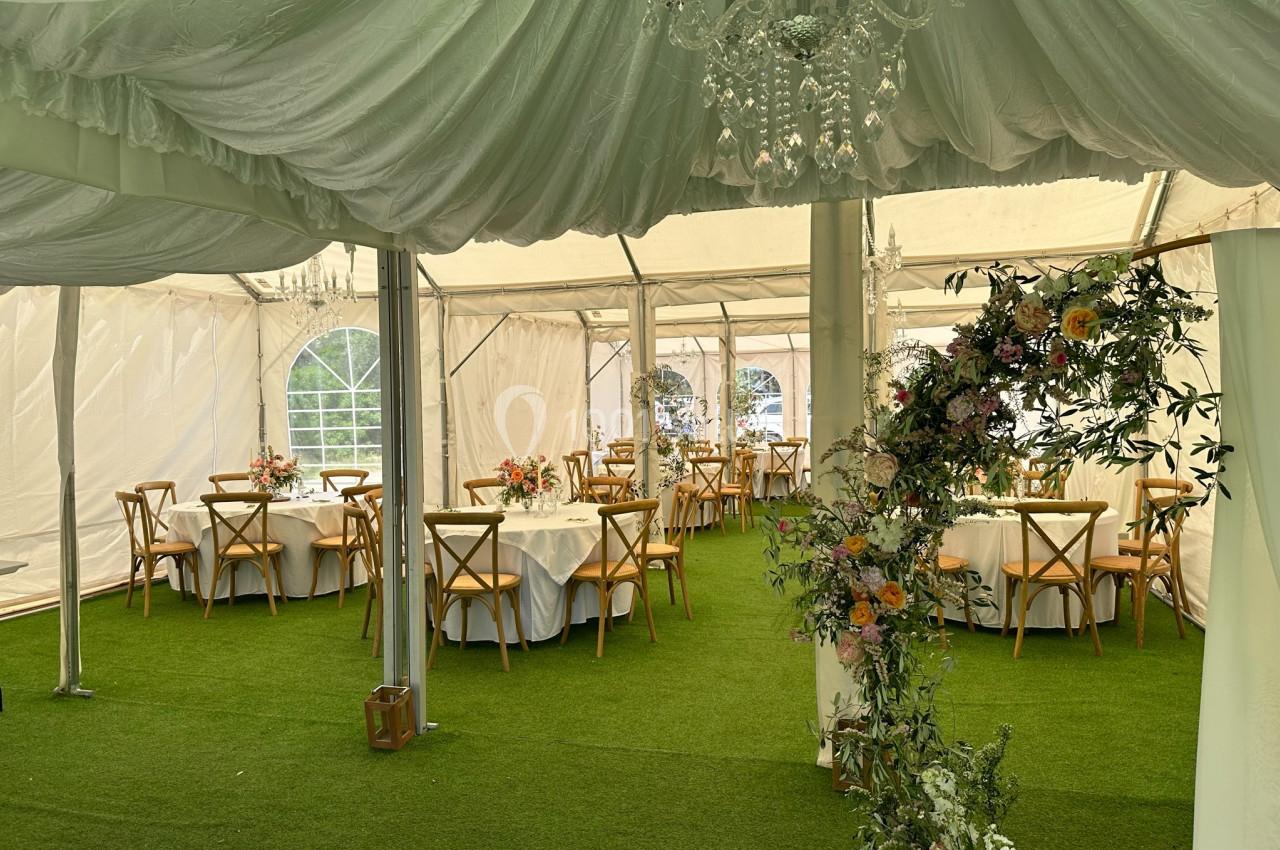 Intérieur d'une tente de réception avec tables rondes décorées et arche florale sur un sol en gazon artificiel.