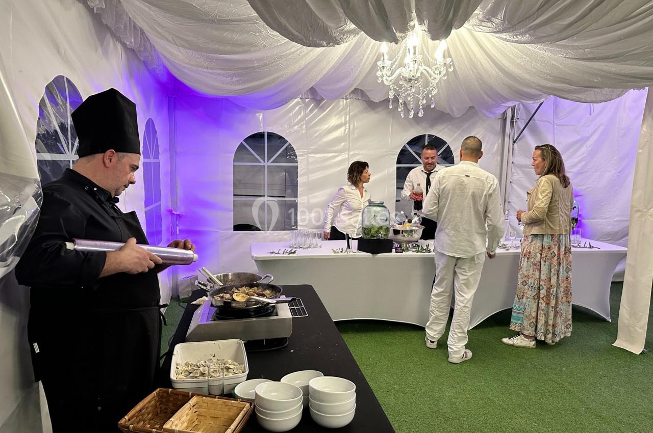 Un chef cuisine sous une tente éclairée, tandis que des invités discutent près d'une table de buffet.