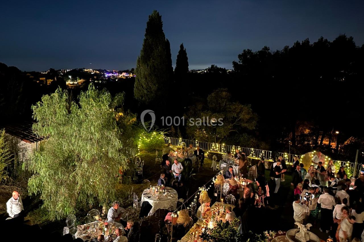 Vue nocturne d'une fête en plein air avec des invités rassemblés autour de tables, entourés de guirlandes lumineuses et de…