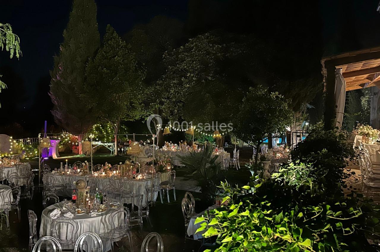 Tables décorées pour un événement en soirée dans un jardin éclairé par des guirlandes lumineuses.