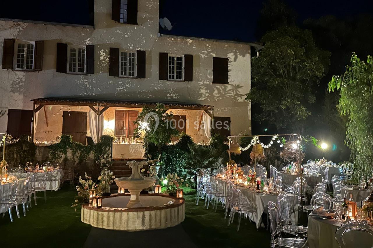Jardin éclairé de nuit avec tables décorées pour un événement, devant une grande maison aux volets ouverts.