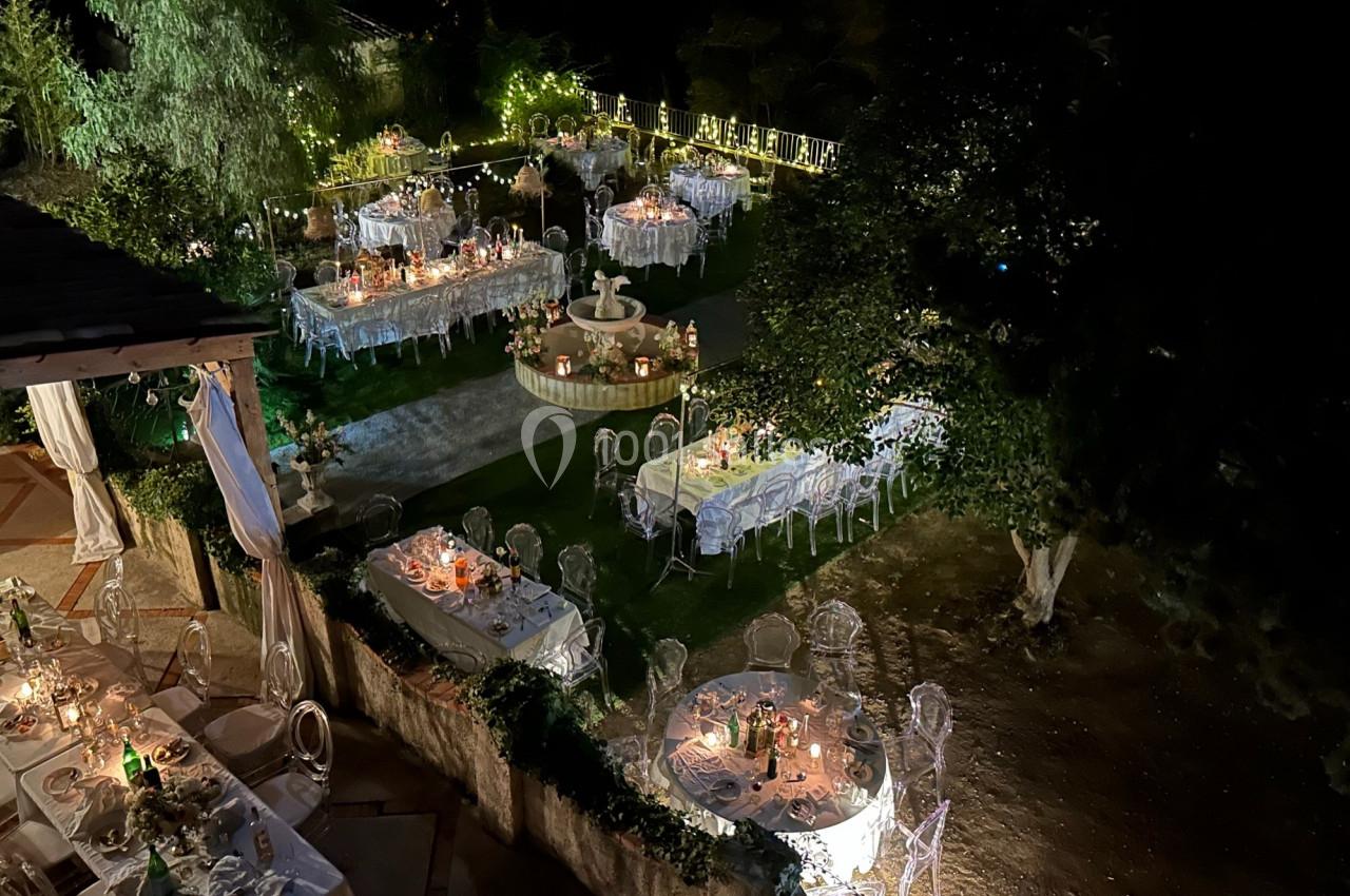 Vue nocturne d'un jardin éclairé avec des tables décorées pour un événement en plein air, entouré de verdure.