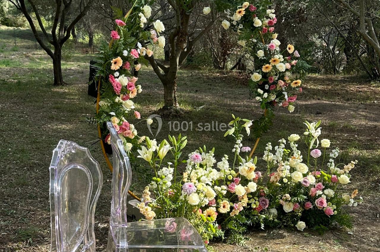 Deux chaises transparentes devant une arche florale circulaire décorée de fleurs colorées dans un jardin boisé.