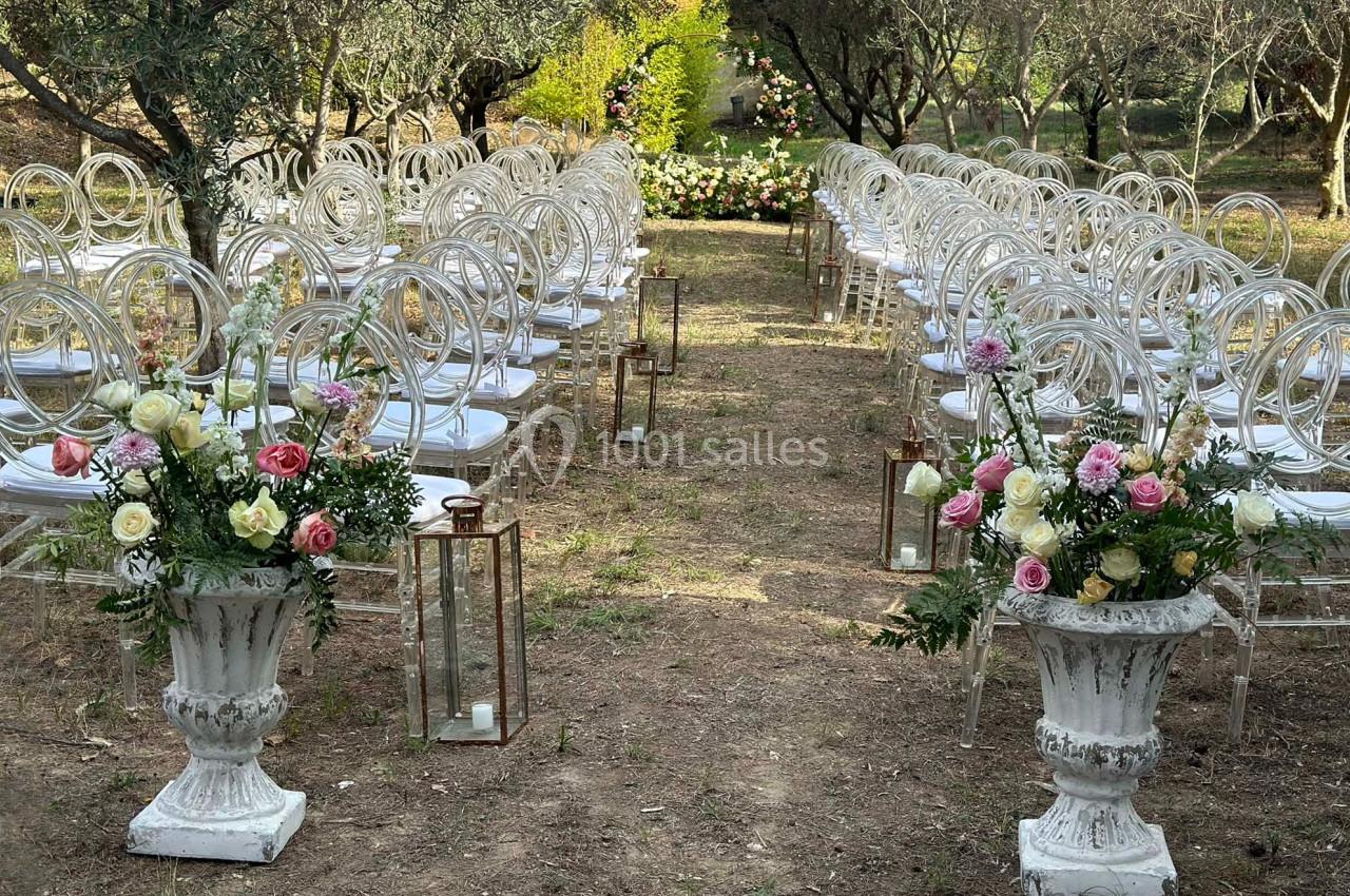 Allée centrale d'une cérémonie en plein air, bordée de chaises transparentes, lanternes et compositions florales.