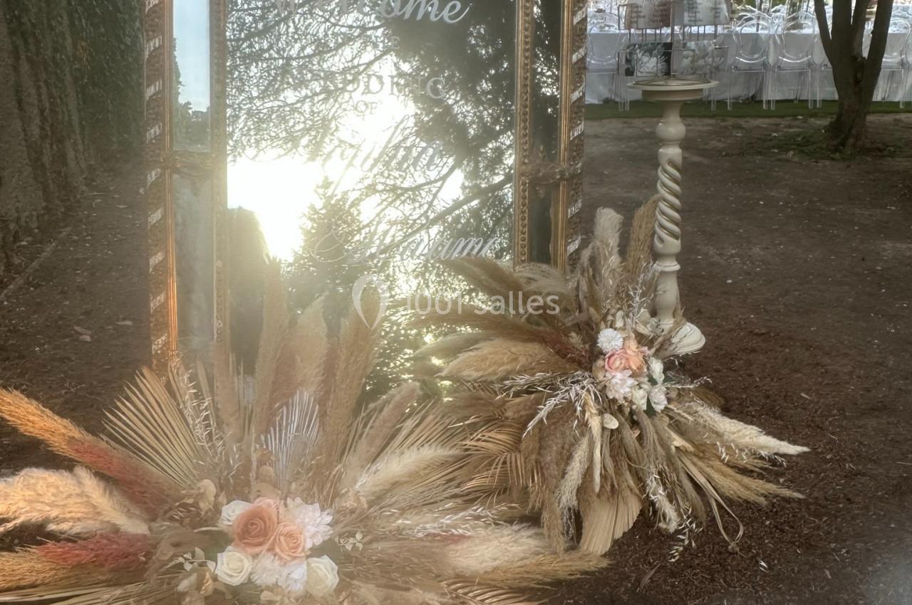 Grand miroir décoratif entouré de compositions florales séchées, placé en extérieur près d'un arbre.