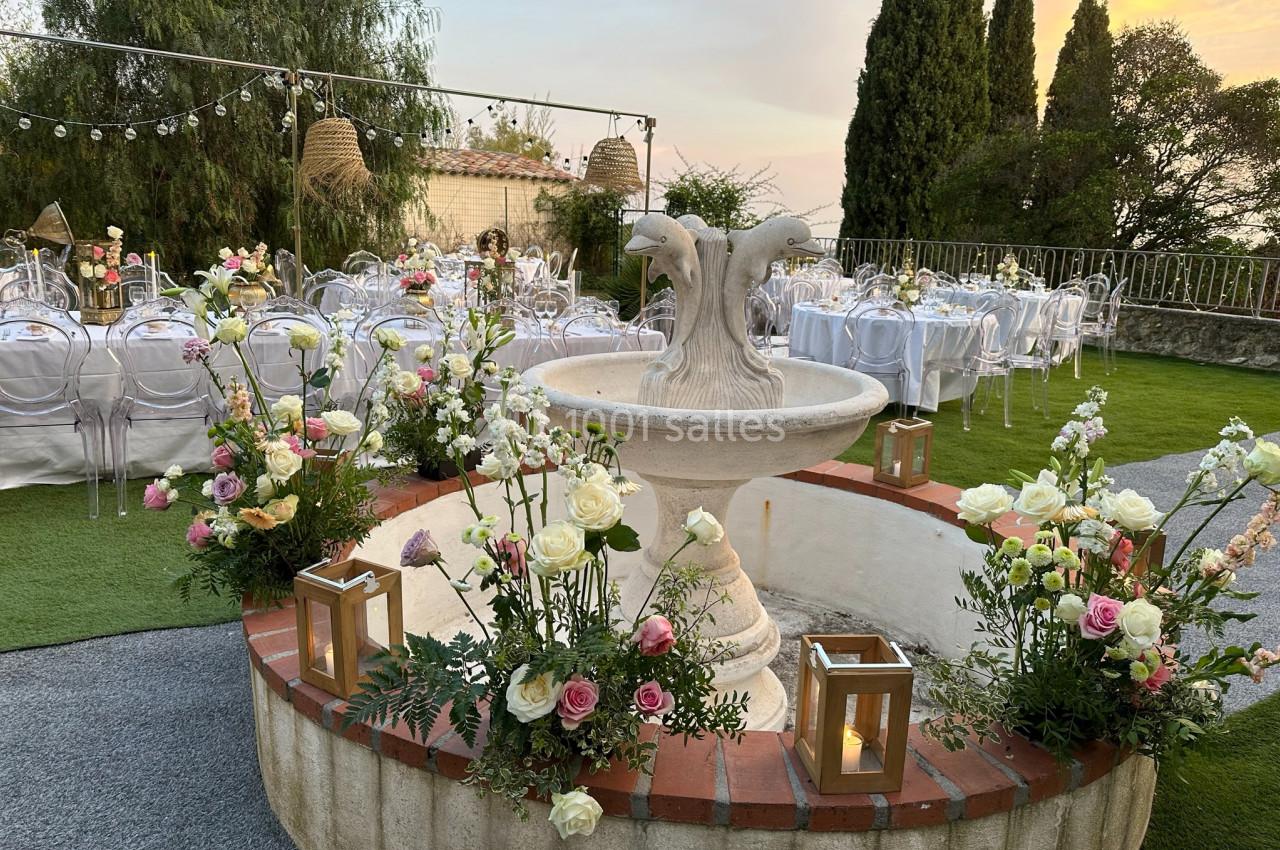 Fontaine décorée de fleurs et de lanternes au centre d'un jardin aménagé pour un événement avec des tables dressées.