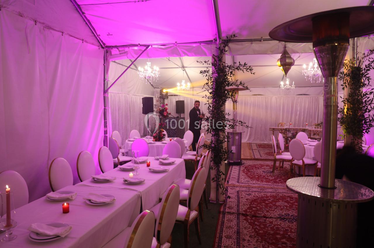 Salle de réception sous tente avec tables dressées, éclairage rose, chandeliers et décorations végétales.