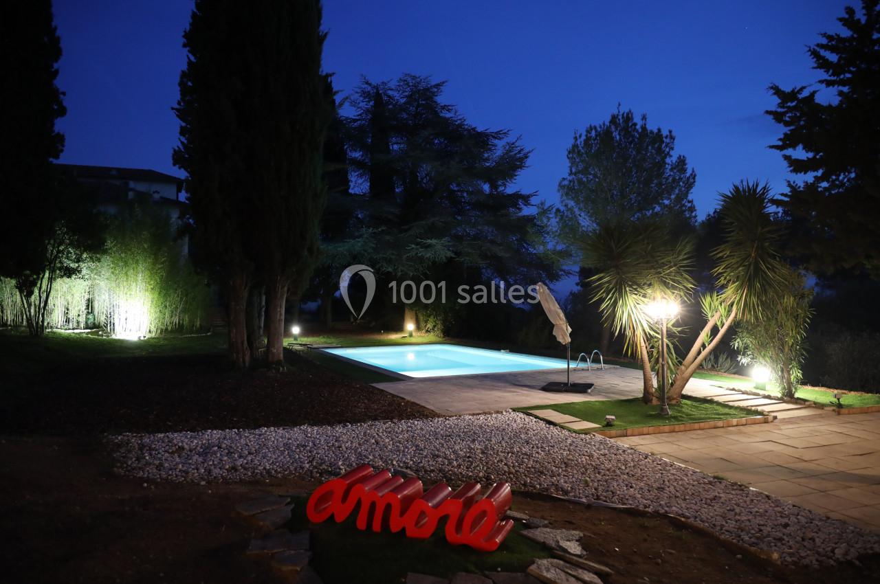 Jardin éclairé de nuit avec piscine illuminée, arbres environnants et sculpture rouge au premier plan.