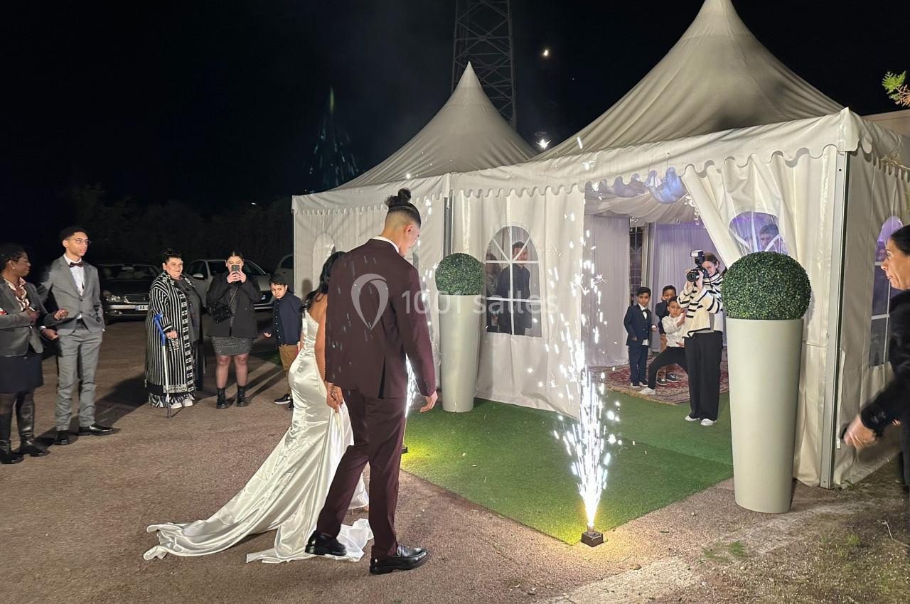 Un couple en tenue de mariage entre sous une tente éclairée, entouré de convives et de feux d'artifice au sol.