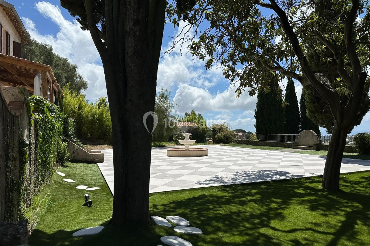 Jardin avec pelouse, arbres, fontaine centrale sur sol pavé et vue dégagée sous un ciel partiellement nuageux.