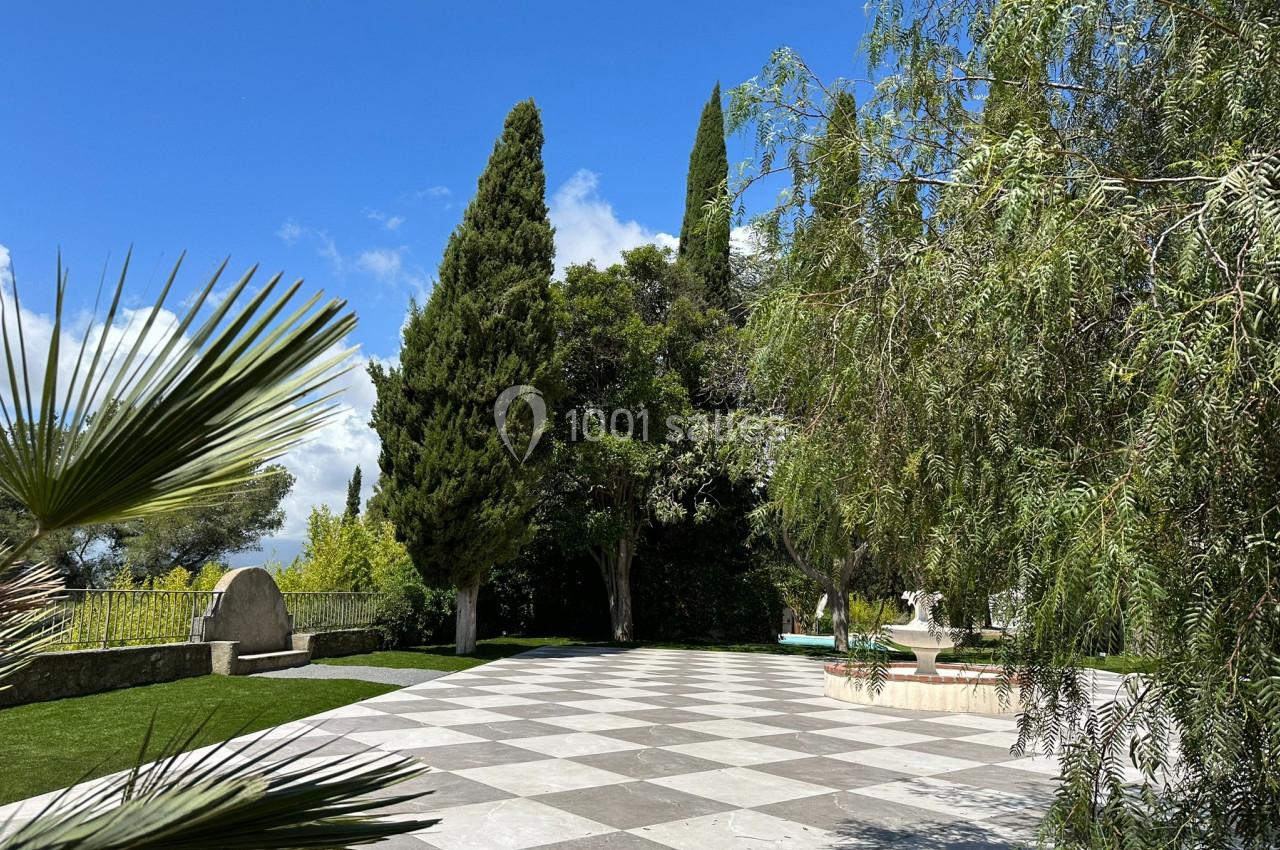 Terrasse pavée entourée de verdure avec des arbres, un ciel bleu et une fontaine en arrière-plan.