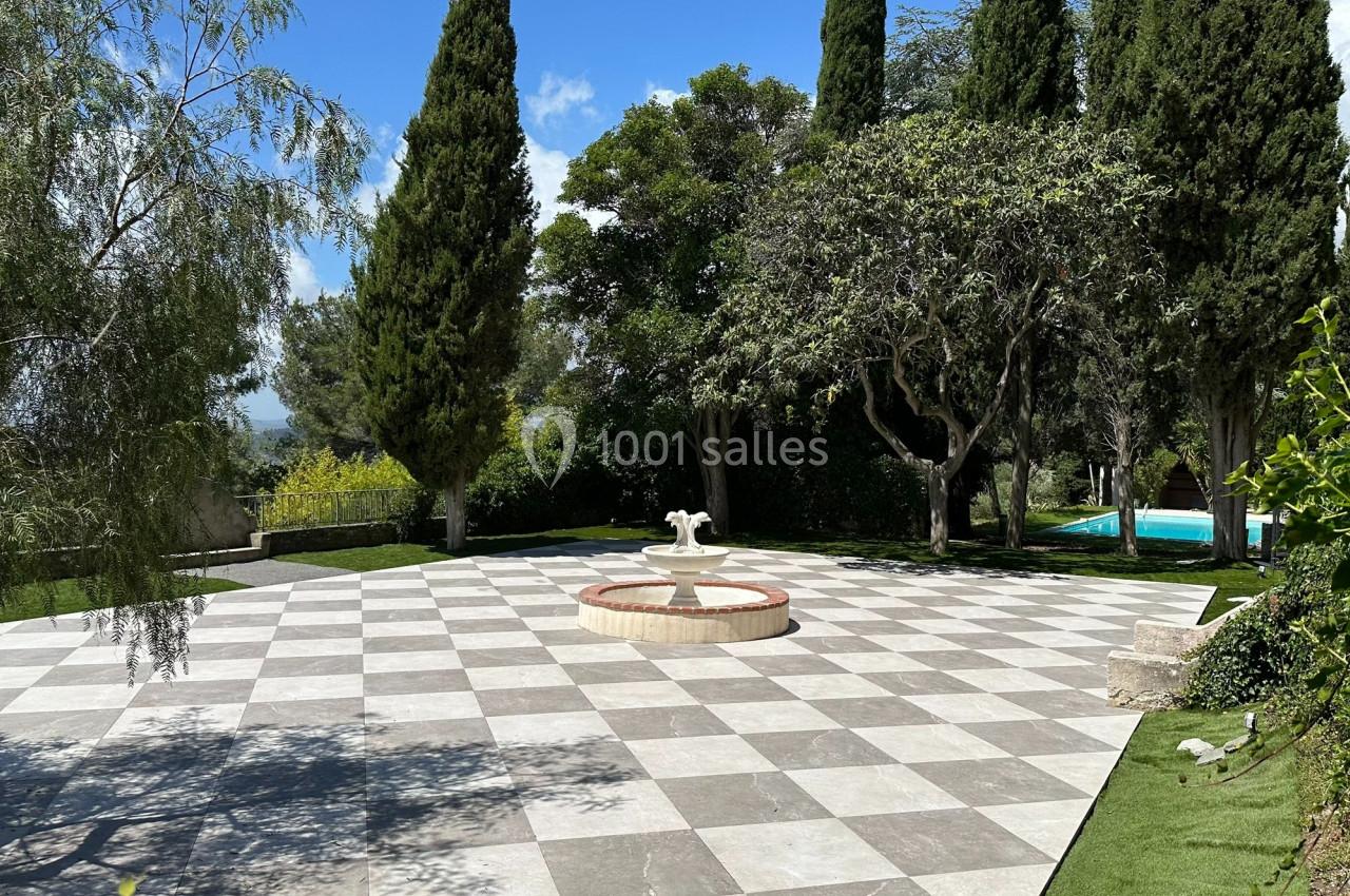 Cour pavée avec une fontaine centrale, entourée d'arbres et de verdure, avec une piscine visible en arrière-plan.