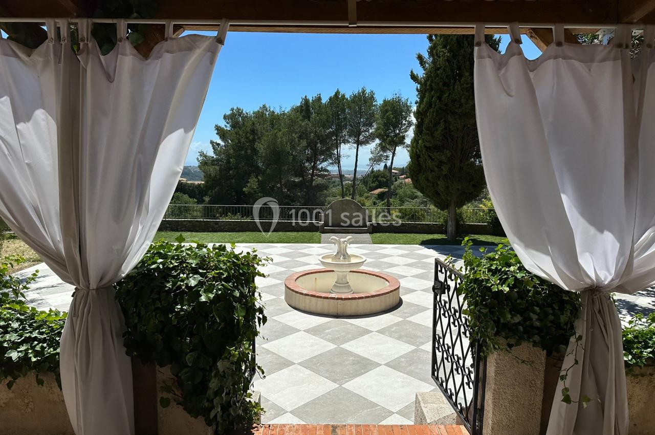 Vue sur une terrasse avec rideaux blancs ouverts, une fontaine centrale et un jardin arboré en arrière-plan.