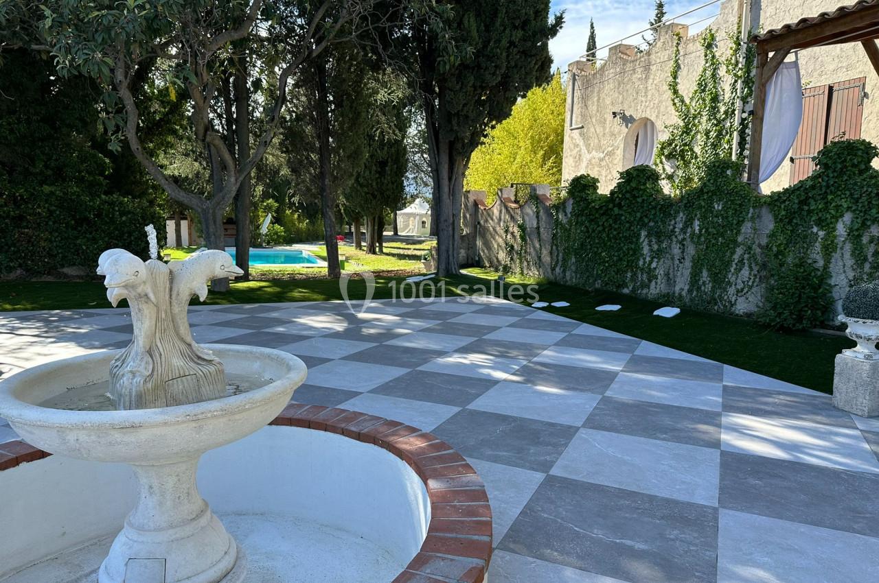 Fontaine en pierre au premier plan sur une terrasse carrelée, avec jardin arboré et piscine en arrière-plan.