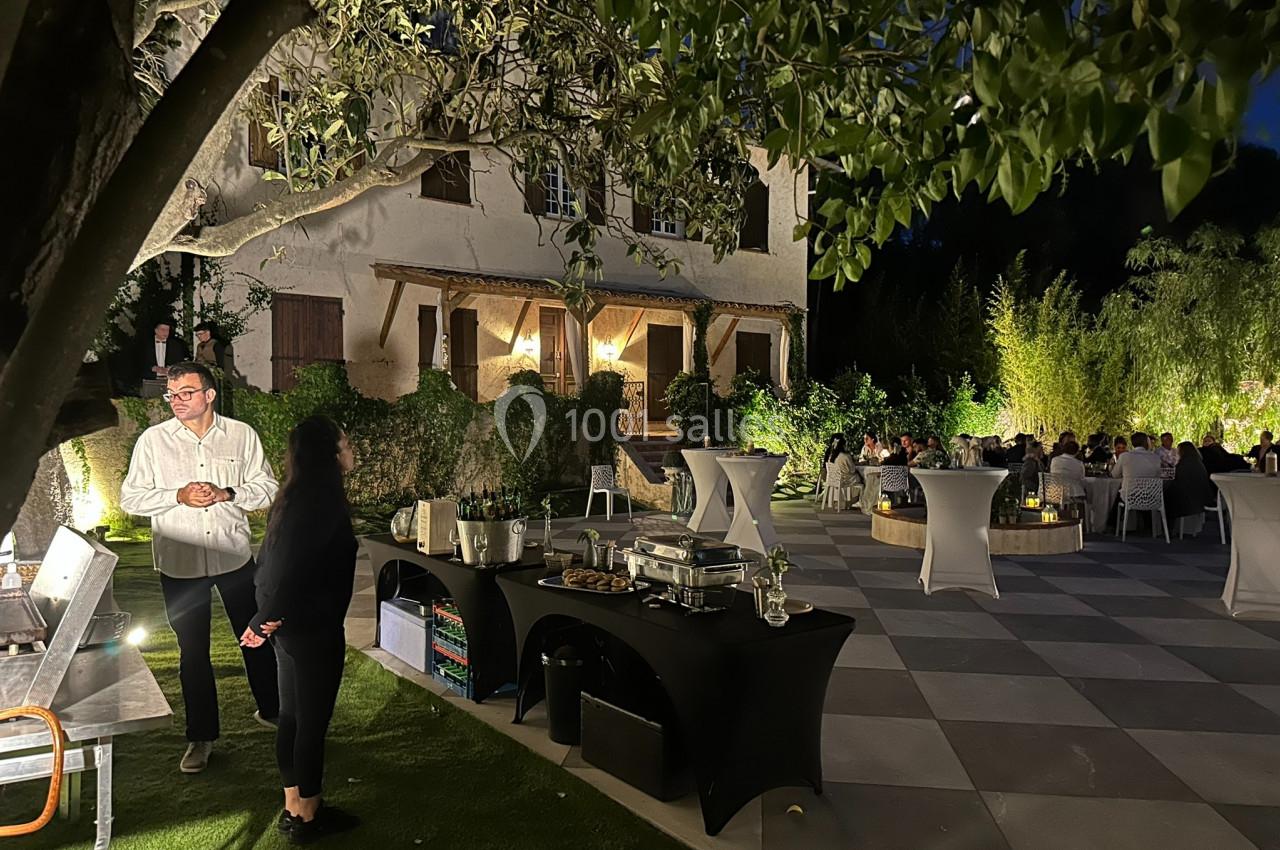 Terrasse éclairée en soirée avec des tables dressées, des invités assis et un buffet sous un arbre.