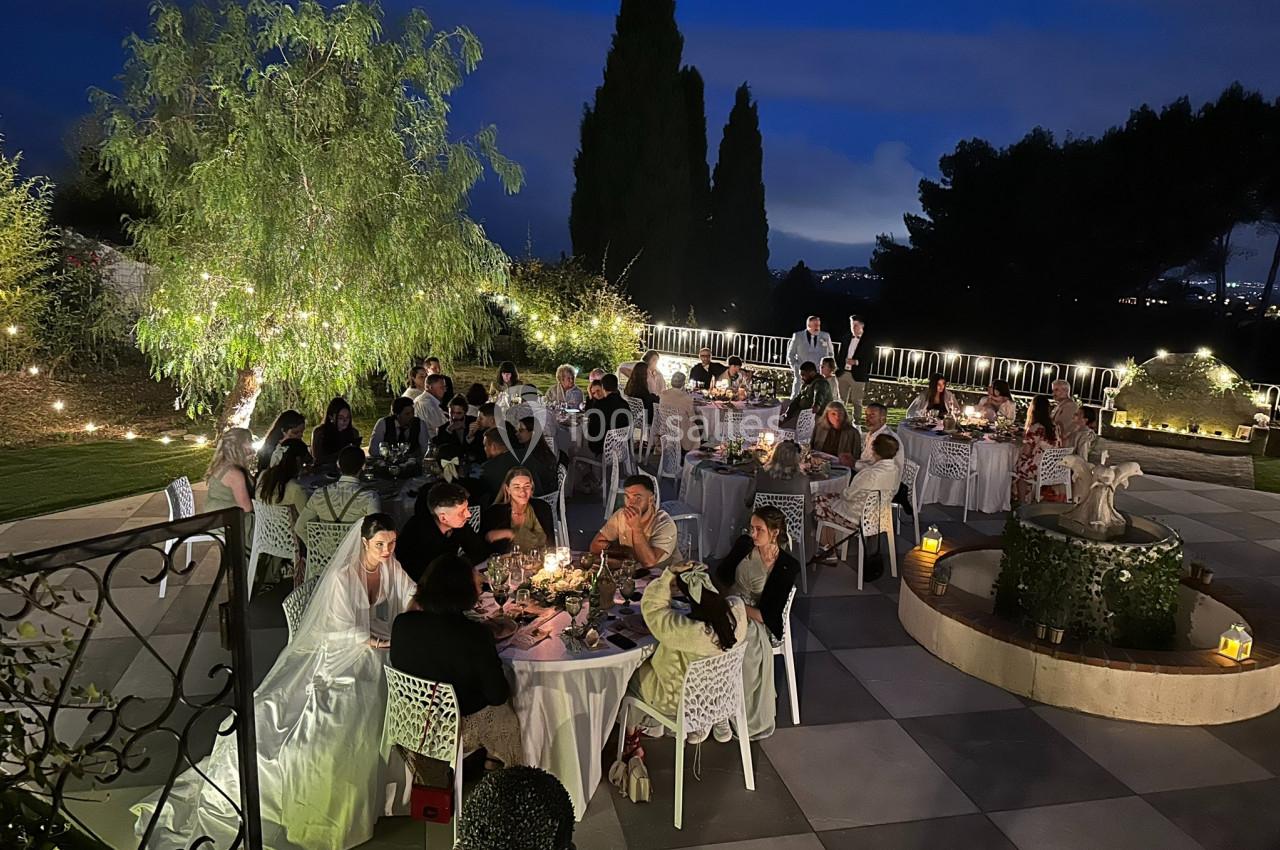 Groupe de personnes dînant en extérieur sur une terrasse éclairée, entourée de végétation, en soirée.