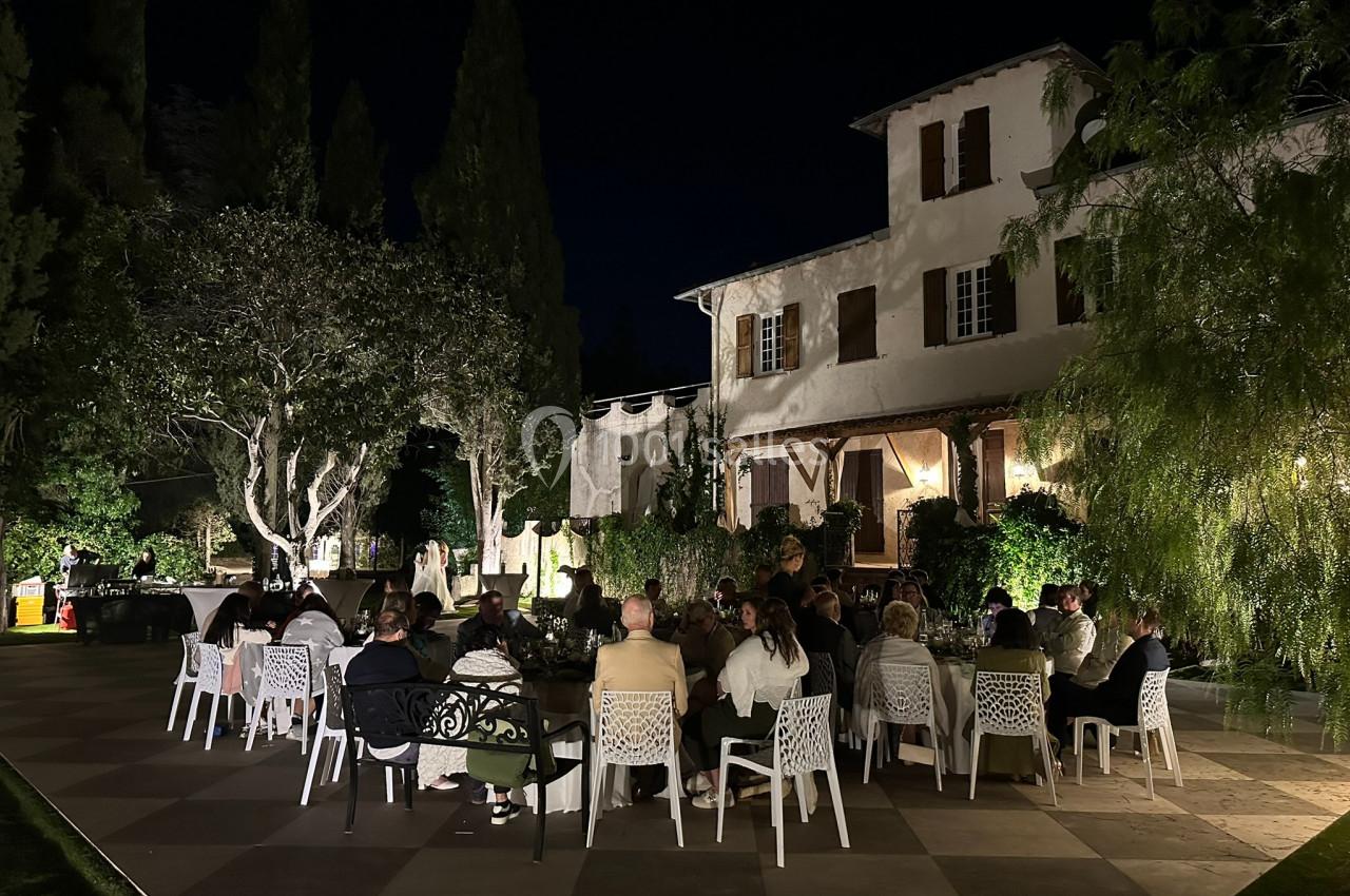 Groupe de personnes dînant en plein air dans un jardin éclairé, devant une grande maison de style traditionnel.