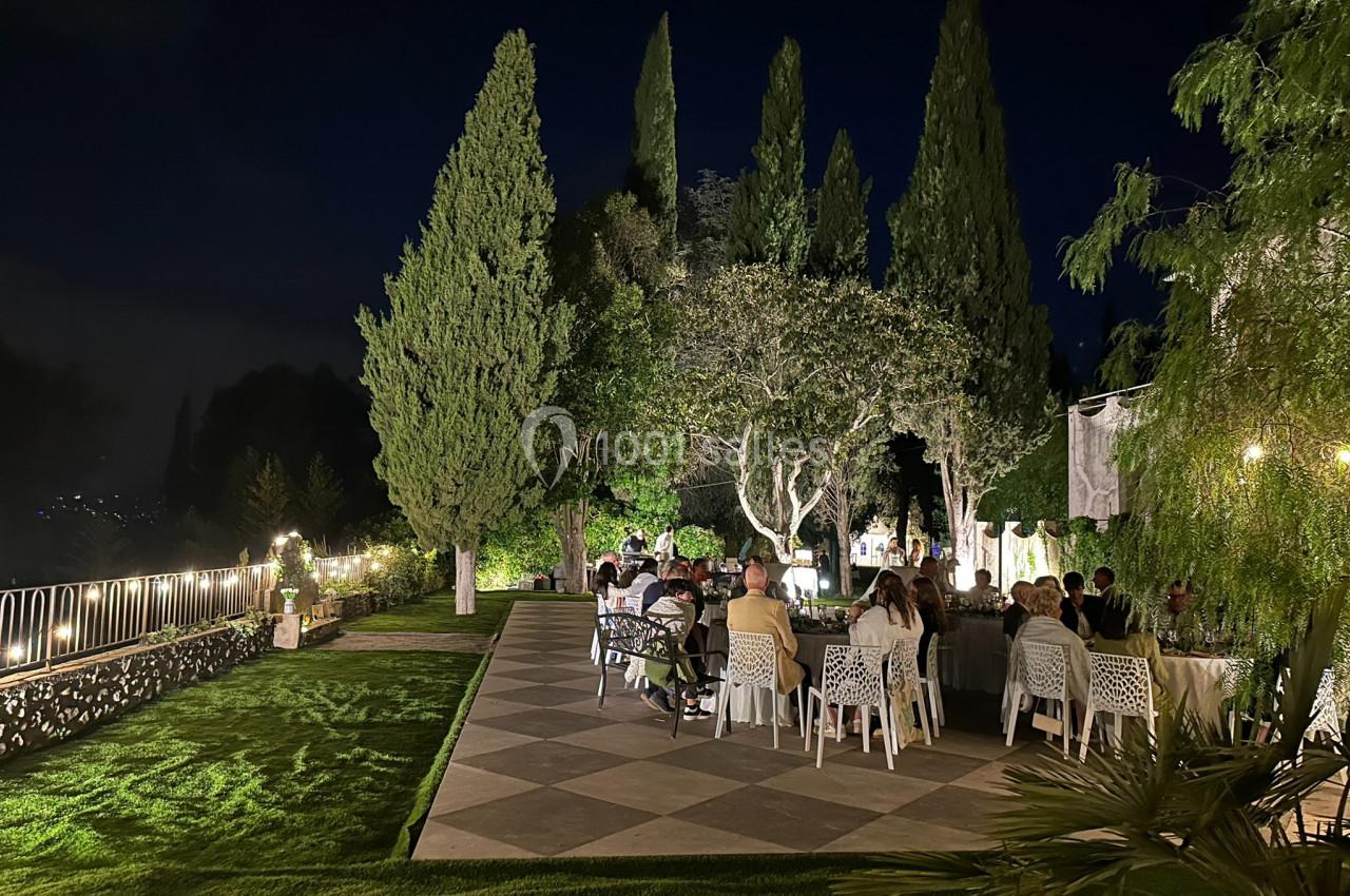 Groupe de personnes dînant en extérieur dans un jardin éclairé, entouré d'arbres et de pelouse, en soirée.