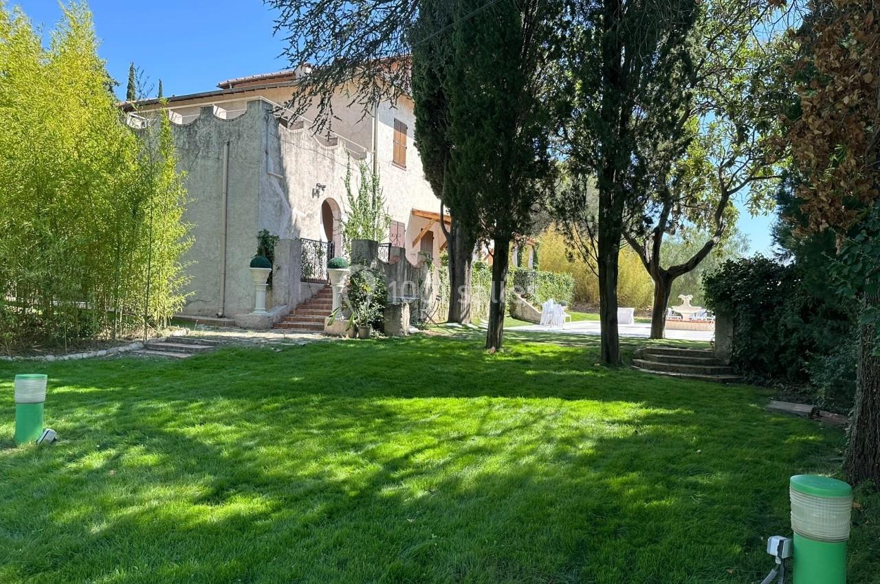 Jardin verdoyant avec pelouse, arbres et une maison en pierre beige avec des volets en bois en arrière-plan.