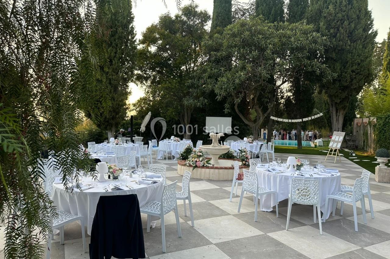 Tables rondes dressées avec nappes blanches dans un jardin extérieur, entouré d'arbres et décoré pour un événement.