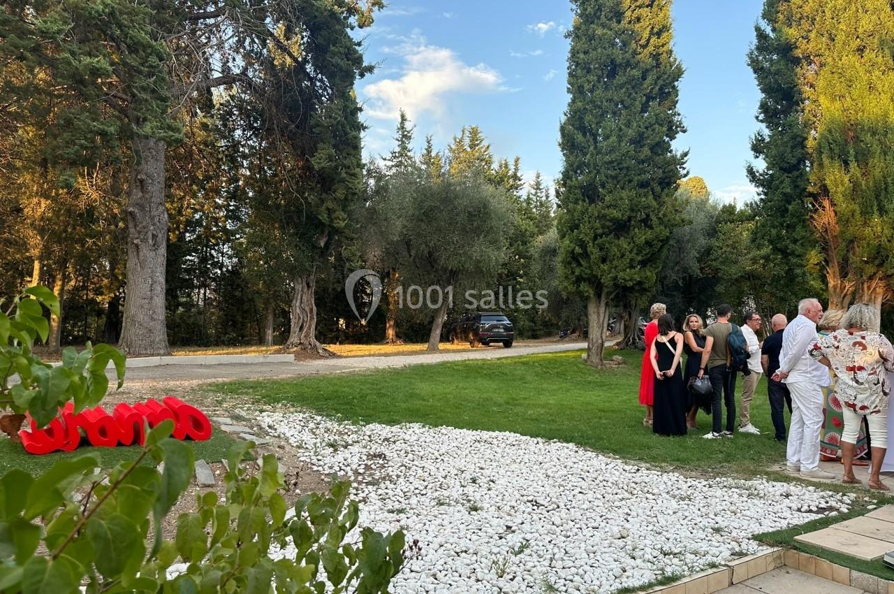 Un groupe de personnes discute dans un jardin bordé d'arbres, avec une allée et une voiture en arrière-plan.
