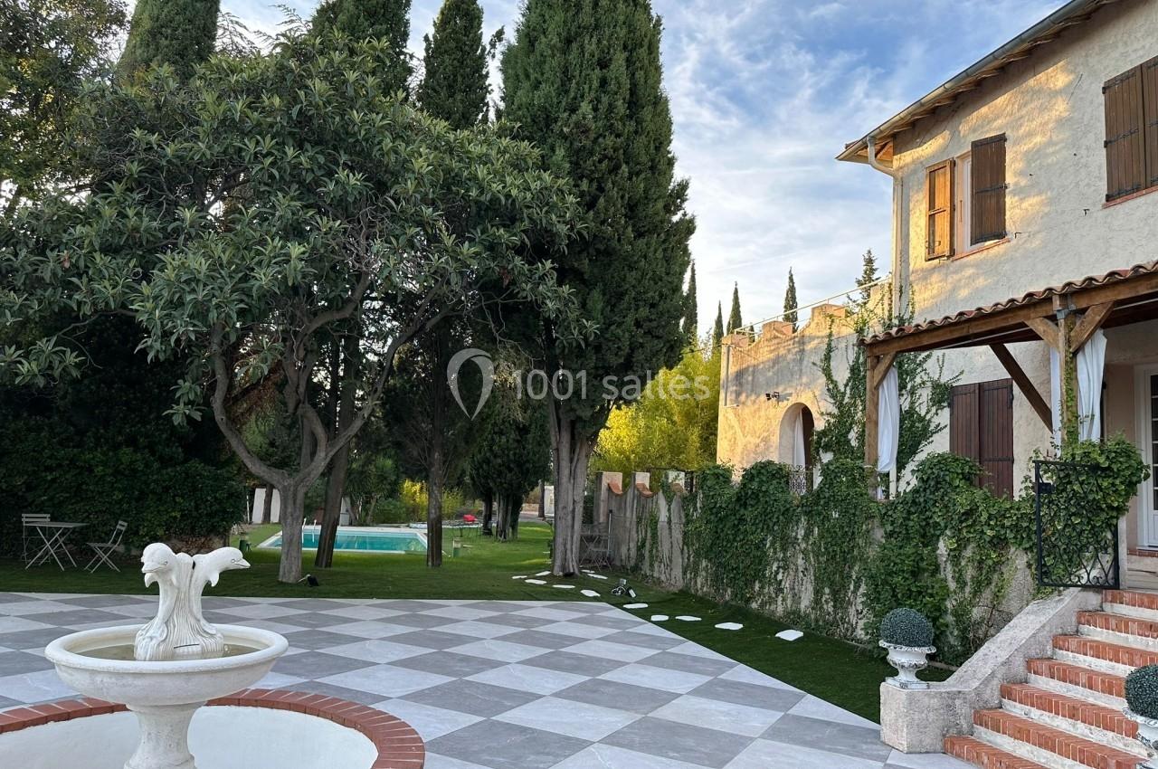 Cour pavée avec fontaine, maison en pierre aux volets en bois, jardin arboré et piscine en arrière-plan.
