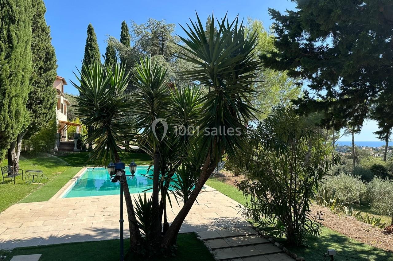 Vue d'un jardin avec piscine entourée de végétation, arbres et plantes, sous un ciel bleu ensoleillé.