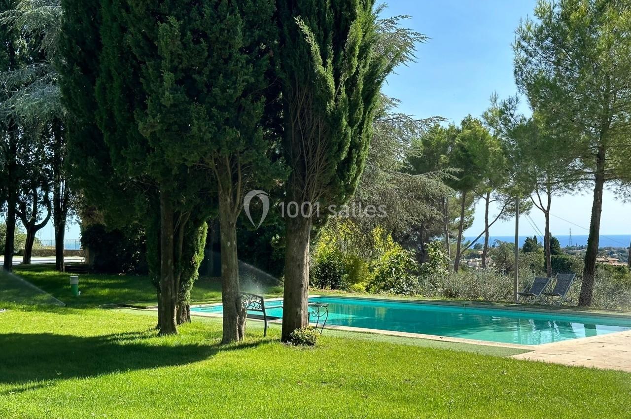 Piscine extérieure entourée de pelouse, arbres et chaises, avec vue sur un paysage ensoleillé et des arbres en arrière-plan.