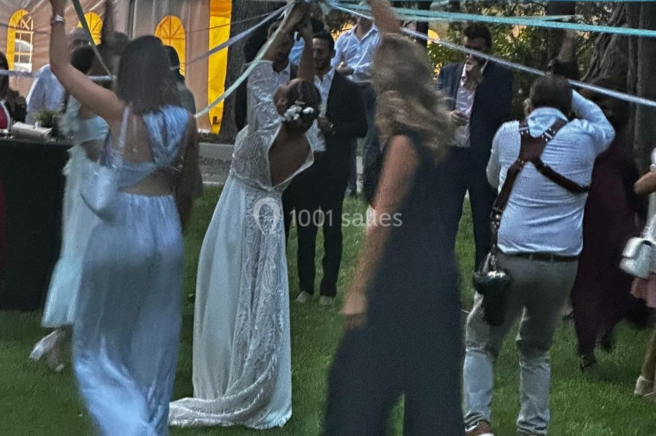 Des invités dansent en plein air lors d'une fête, avec une mariée en robe blanche au centre tenant des rubans.