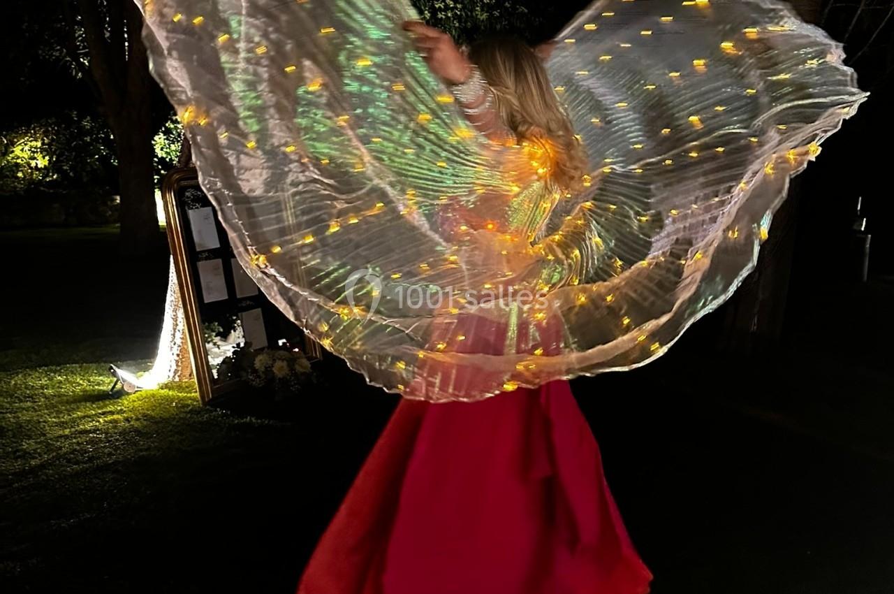 Une personne en robe rouge danse de nuit avec des ailes lumineuses scintillantes dans un jardin sombre.