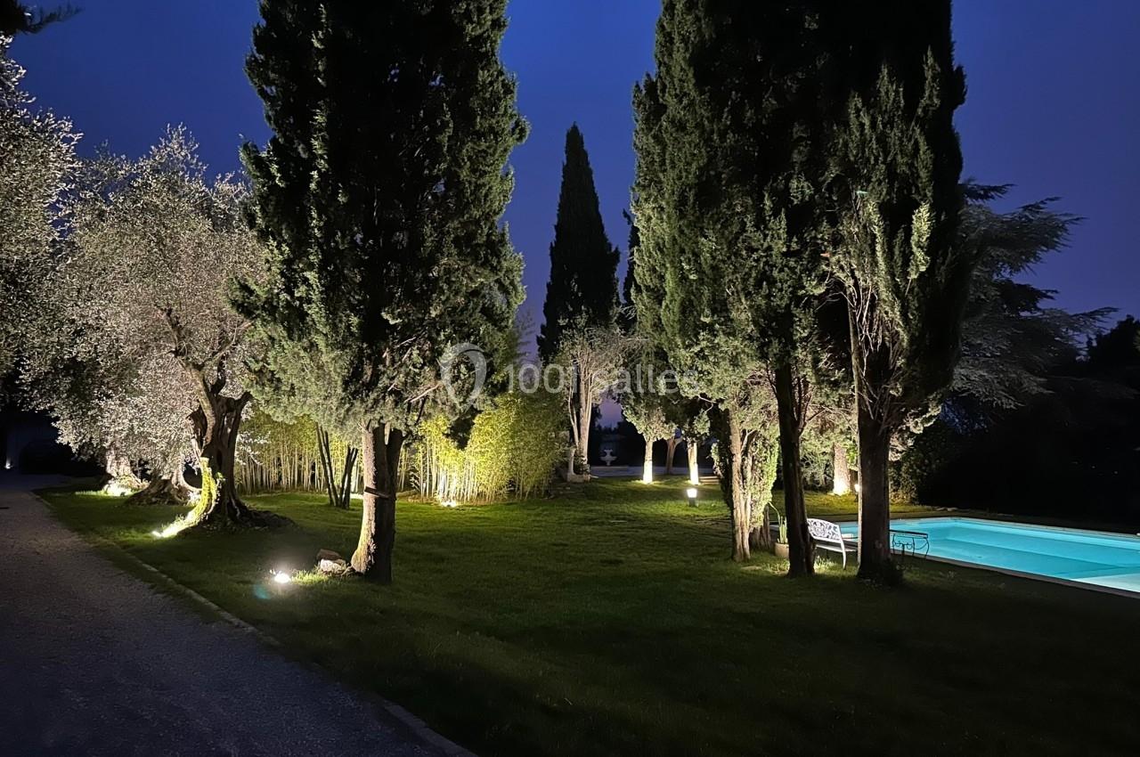 Jardin éclairé de nuit avec des arbres, une pelouse et une piscine illuminée à droite.