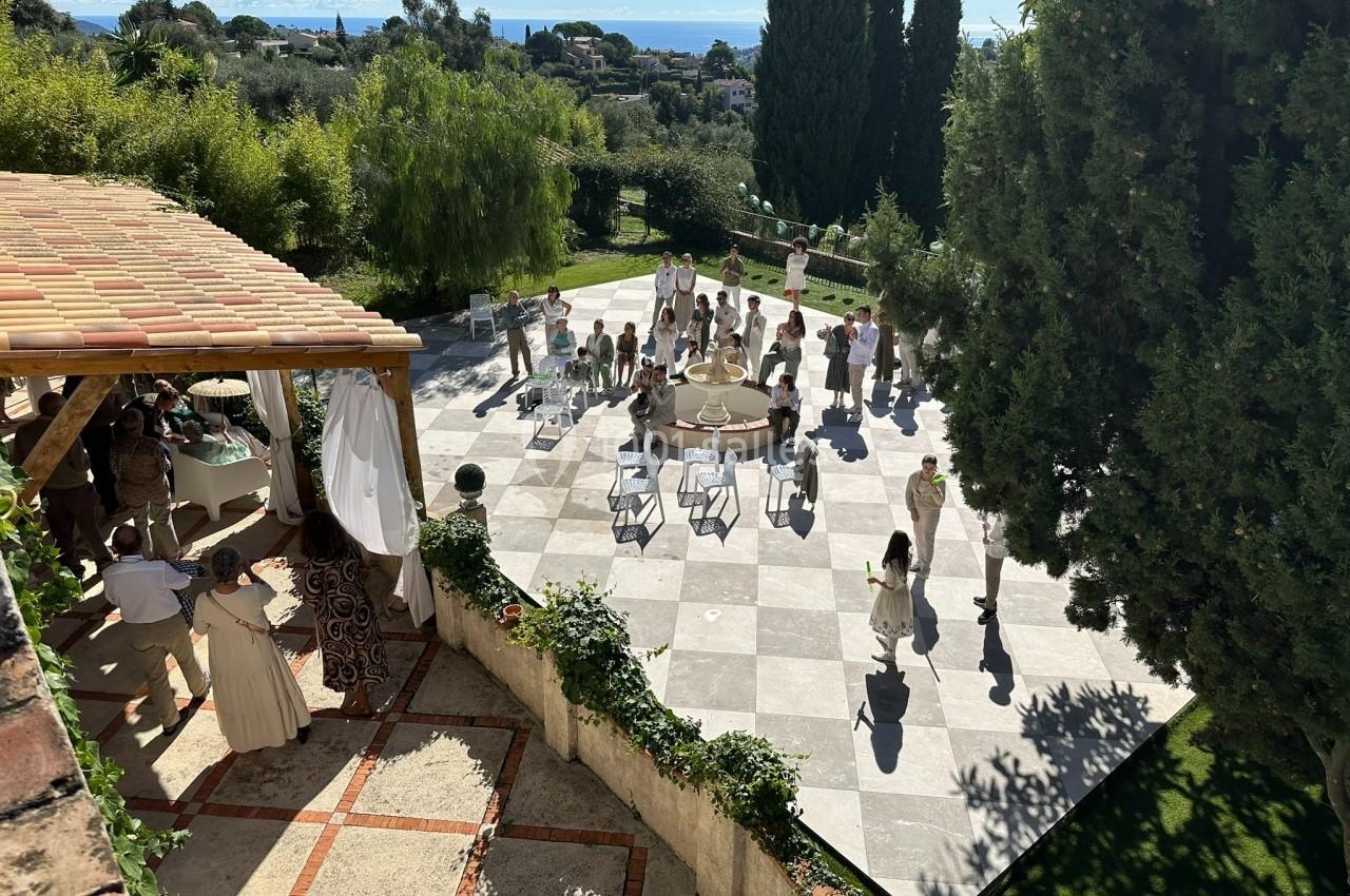 Groupe de personnes rassemblées sur une terrasse ensoleillée entourée de verdure, avec vue sur la mer au loin.